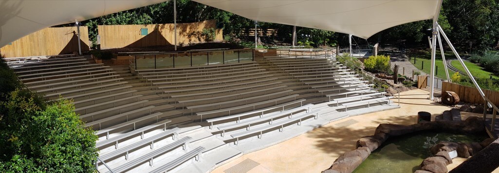 An outdoor amphitheater with rows of long benches arranged in tiered seating, shaded by a large white canopy. Steps lead up to each row, and the area is surrounded by lush greenery and plants.