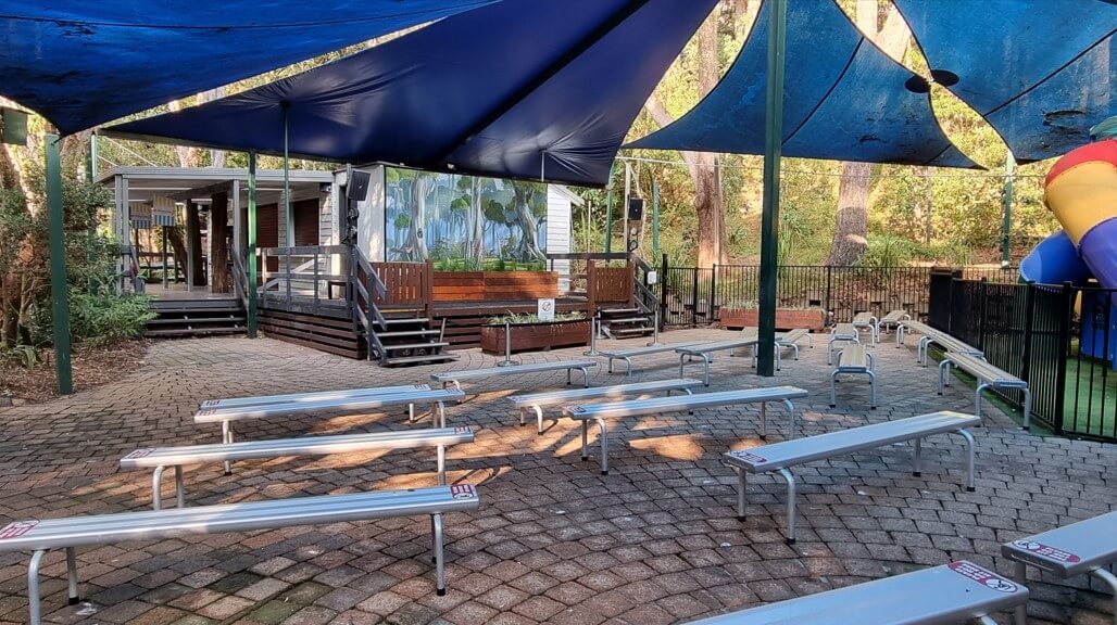 An outdoor area with multiple rows of metal benches on a paved surface, shaded by large blue shade sails.