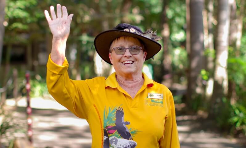 An older woman in a yellow shirt and hat smiles and waves outdoors on a sunny day. She is wearing glasses and a volunteer badge.