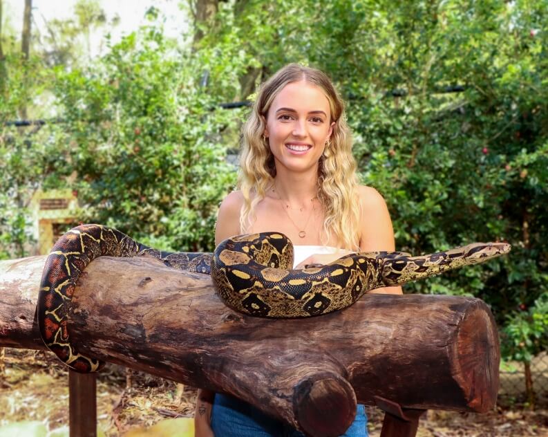 A woman with long blonde hair stands outdoors behind a log, holding a large snake resting on the log in front of her. Trees and greenery are in the background.