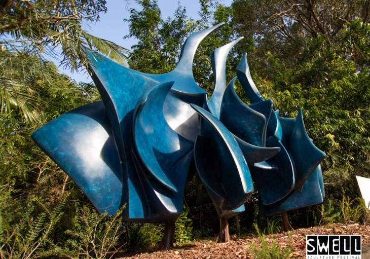 Abstract blue sculpture with curved, pointed shapes displayed outdoors among green trees and plants; SWELL Sculpture Festival logo at bottom right.
