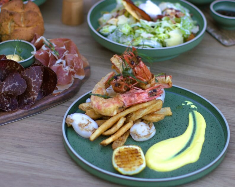 A beautifully arranged plate featuring crispy fried shrimp, golden French fries, grilled scallops, a slice of grilled lemon, and a vibrant yellow sauce.