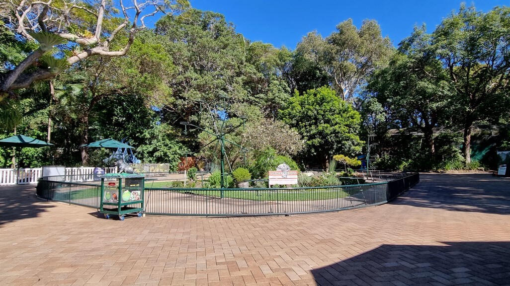 A landscaped garden enclosed by a black metal fence, featuring lush greenery, trees, and a sign that reads 'Welcome to the Garden'.