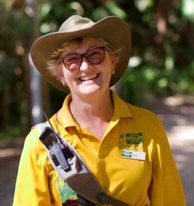Smiling woman in a yellow polo shirt and a brown hat, wearing sunglasses and carrying a radio, likely a tour guide.