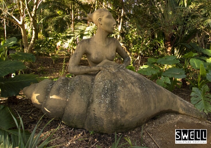 A large outdoor sculpture of a woman with a shell-shaped lower body is surrounded by tropical plants; the SWELL Sculpture Festival logo appears in the corner.