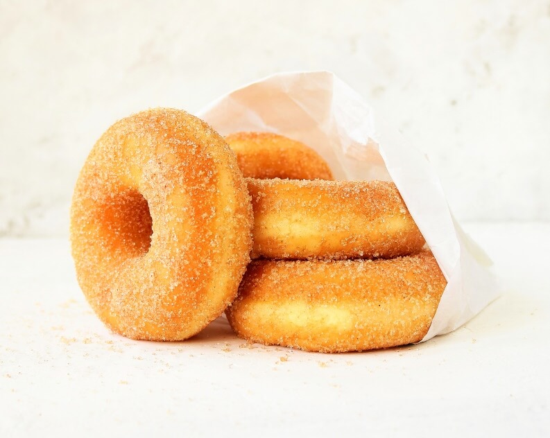 Four golden-brown donuts covered in granulated sugar, with three partially tucked inside a white paper bag and one resting in front.
