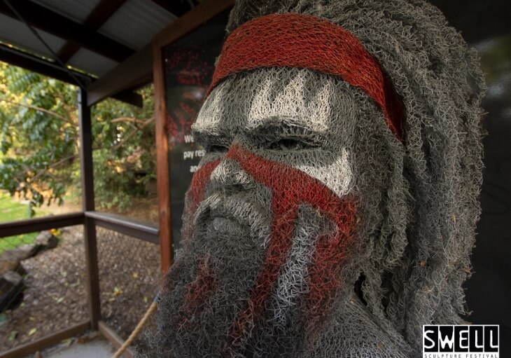 A wire mesh sculpture of a persons head and shoulders with red and white face paint, displayed at the SWELL Sculpture Festival.