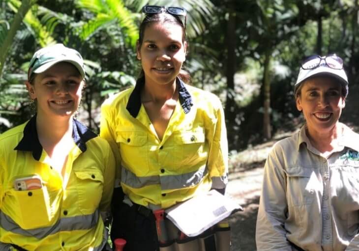 Three women wearing high-visibility work shirts and hats stand outdoors in a forested area, smiling at the camera.