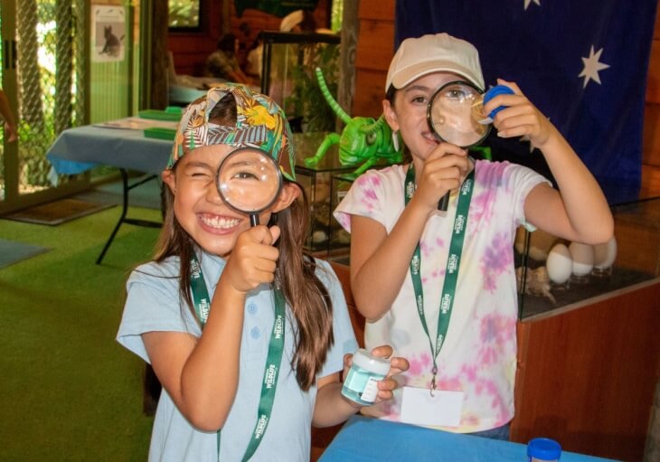 Two children, smiling and holding magnifying glasses, explore insects at a natural history museum.