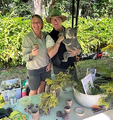 Two people with a koala and surrounded by plants and seedling pots, participate in a nature-based educational event.
