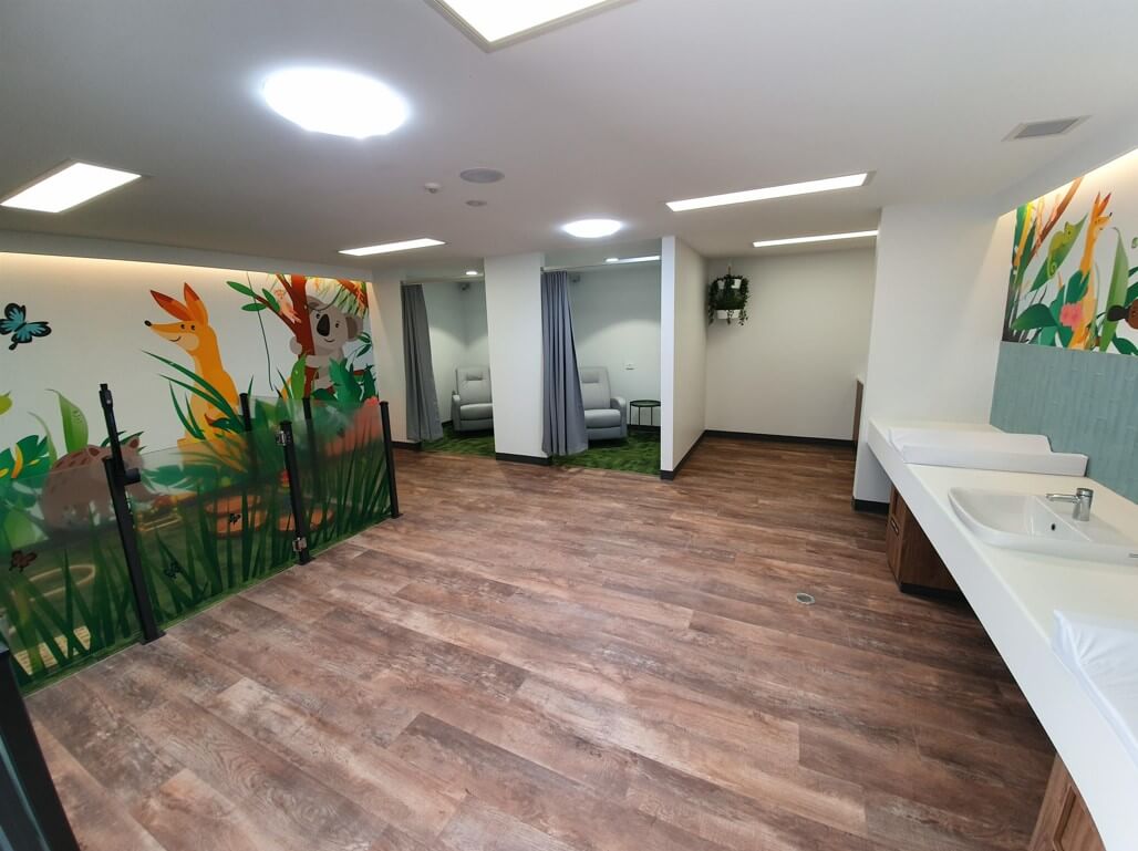 A brightly lit room with wooden flooring and colorful cartoon animal and plant murals on the wall. On the right side, two sinks with white countertops are visible.