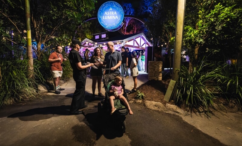 A group of people, including a child in a stroller, stands on a path at night near the illuminated entrance sign for Astra Lumina, surrounded by trees and lighting.