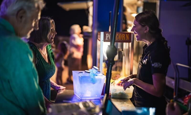A woman behind a counter talks to two people near a glowing ice display and a popcorn machine at an outdoor event at night.