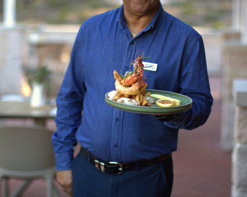 Waiter holding a plate of fried seafood, including shrimp, french fries, and a lemon wedge, outdoors.