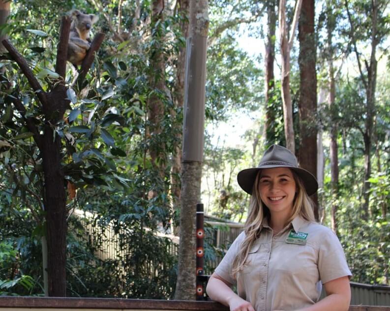 A woman in a khaki uniform and hat stands smiling in front of a fence; a koala sits in a tree behind her amid dense forest.