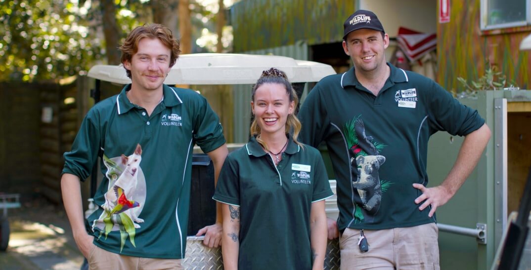 Three zoo volunteers in green shirts with animal prints stand outdoors, smiling at the camera, near a vehicle and a building.