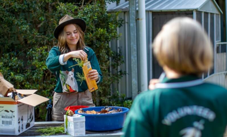 Two people wearing green volunteer shirts work outdoors; one pours liquid into a container with leaves and branches, while the other stands in the foreground facing away.