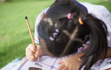 A child sits on a grassy surface, focused on writing in a workbook with a pencil.