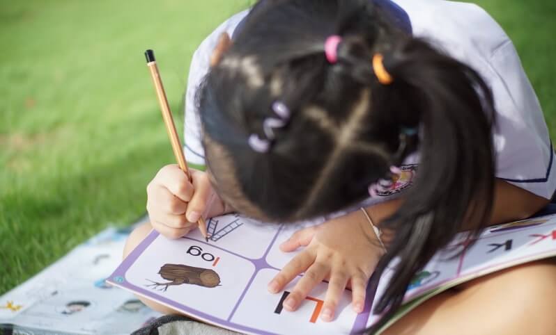 A child sits on a grassy surface, focused on writing in a workbook with a pencil.