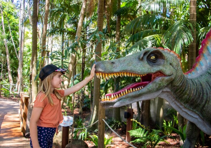 A young girl touches the snout of a large dinosaur statue in an outdoor, tropical-themed setting.