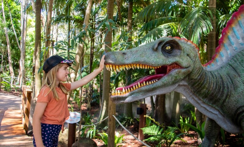 Girl reaching out to touch a large, realistic dinosaur model in a lush, tropical setting.