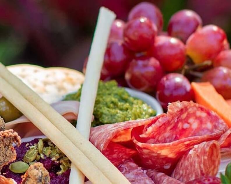 A close-up of a charcuterie board with sliced salami, breadsticks, grapes, crackers, pesto, olives, and vegetables.