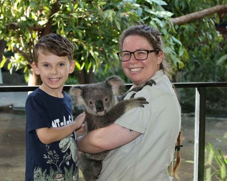 A smiling child stands next to an adult holding a koala in an outdoor setting with green foliage in the background.