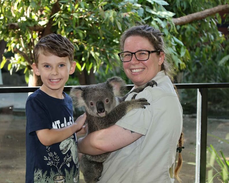 A smiling child stands next to an adult holding a koala in an outdoor setting with green foliage in the background.