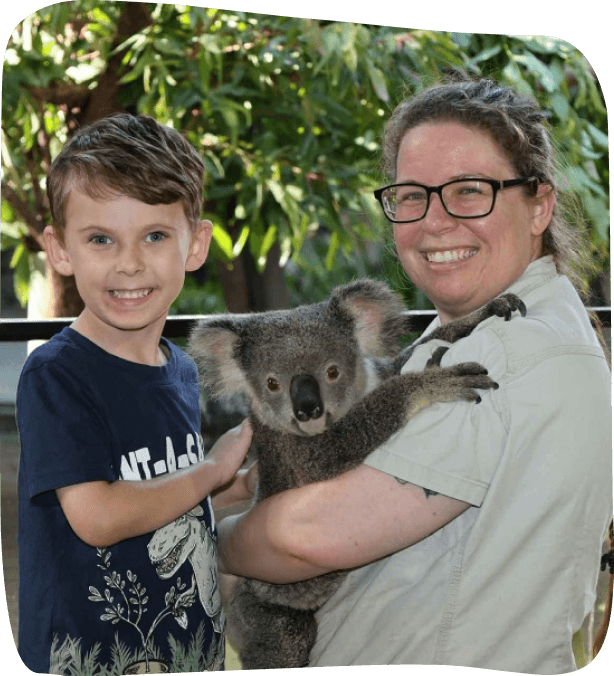 A woman holds a koala while standing next to a smiling young boy. They are outdoors with green foliage in the background.