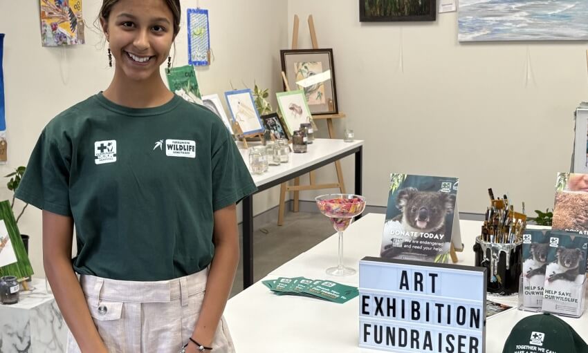 A woman stands in front of a table displaying art and donation materials at an art exhibition fundraiser for wildlife conservation.