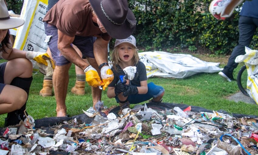 A child and an adult wearing gloves pick up litter from a large pile of trash outdoors, participating in a community cleanup event.