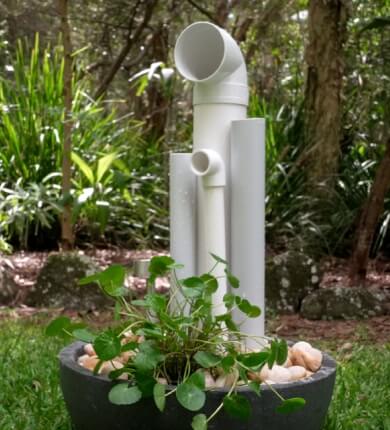 White PVC pipes arranged vertically in a planter with green foliage and rocks, set outdoors on grass with trees in the background.
