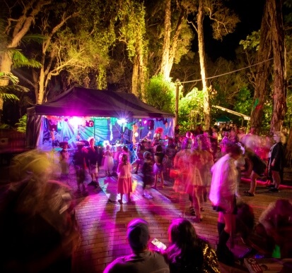 A crowd of people, including children, gathers under colorful lights at an outdoor nighttime event surrounded by tall trees.