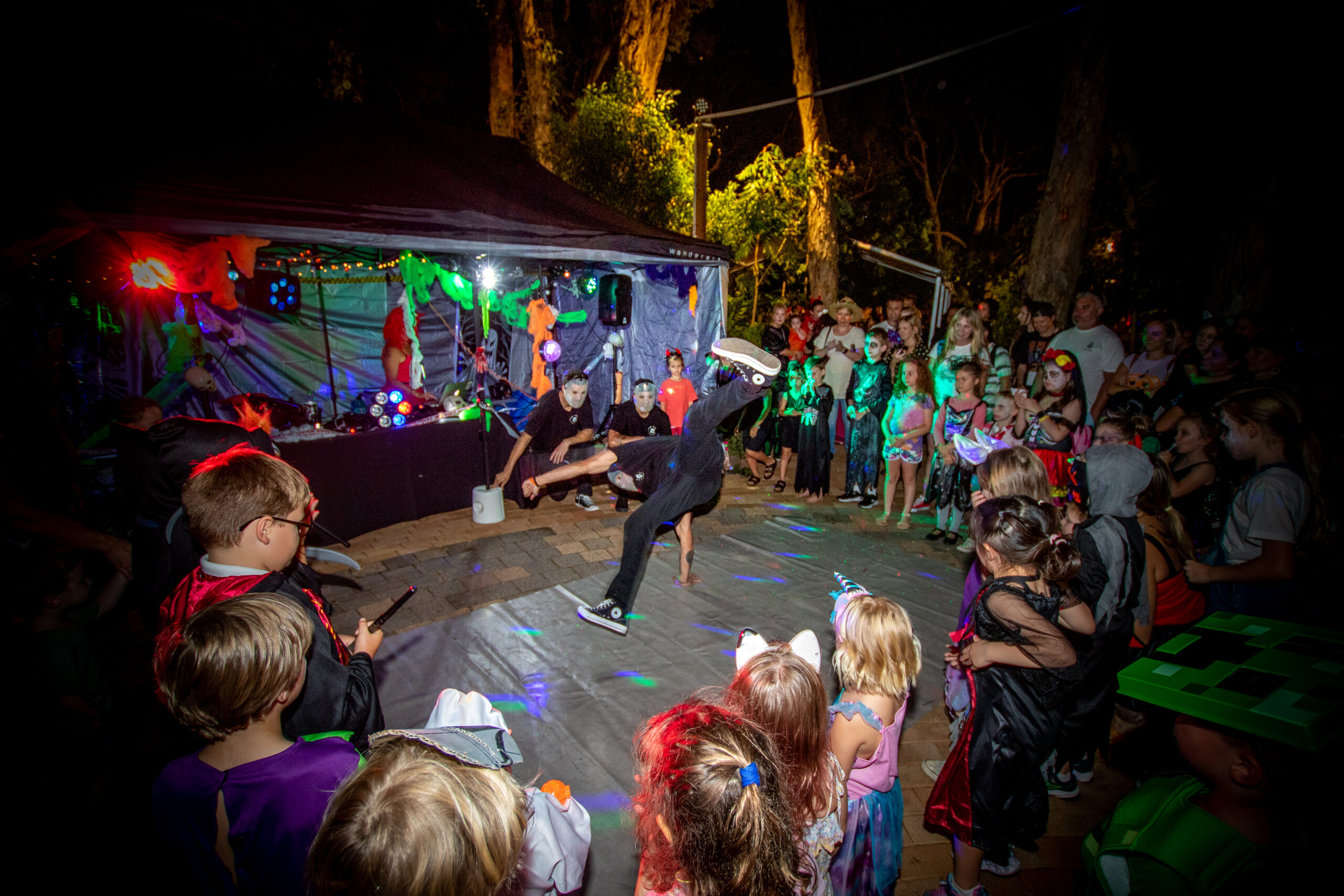 A performer breakdances for a crowd of people surrounding them in a semicircle, in front of a colourful stage at night.
