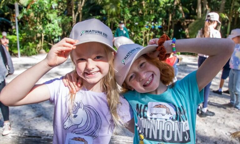 Two smiling children wearing Nature Ed hats and colorful t-shirts pose together outdoors, with other children and greenery visible in the background.