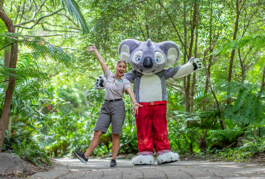 A person in a zoo uniform poses with someone in a large koala costume wearing red pants, surrounded by lush green trees and plants on a stone path.
