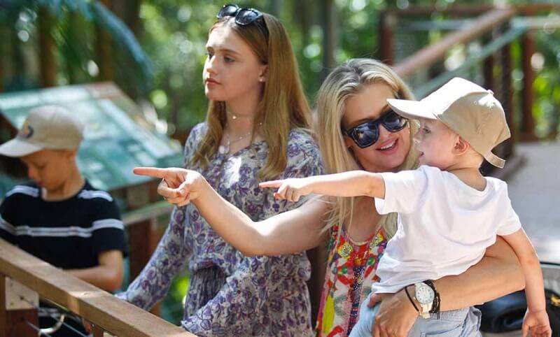A woman holding a young boy points while he also points; a teenage girl and another boy stand nearby in an outdoor, wooded setting.