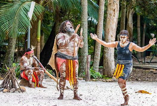 Three Indigenous Australians in traditional attire and body paint perform a cultural dance outdoors, surrounded by trees and natural vegetation.
