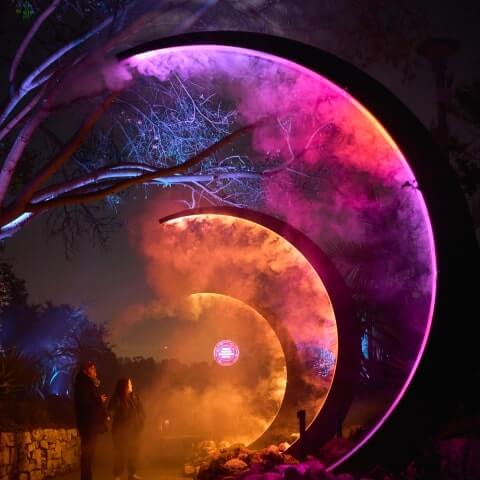 Two people stand under large, illuminated crescent-shaped arches emitting pink and orange light and mist at night, with trees and plants in the background.