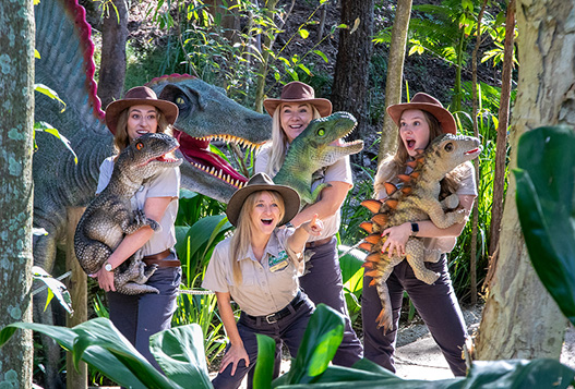 Four women in ranger uniforms pose with dinosaur models and puppets in a lush outdoor setting with trees and greenery.
