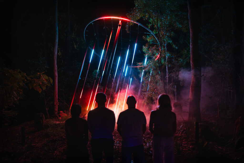 Four people stand on a wooden platform at night, silhouetted against bright orange and blue lights and smoke in the background.