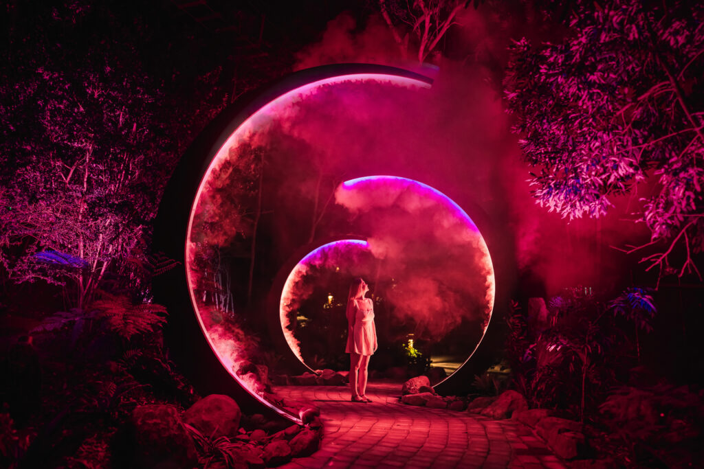 Woman walking through a foggy, red illuminated tunnel of crescent-shaped arches at night, surrounded by trees and colorful lights.