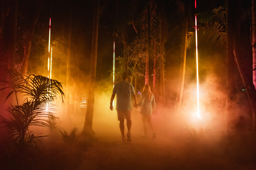 Two people walking hand in hand through a forested area with orange lights and smoke effects amongst large trees