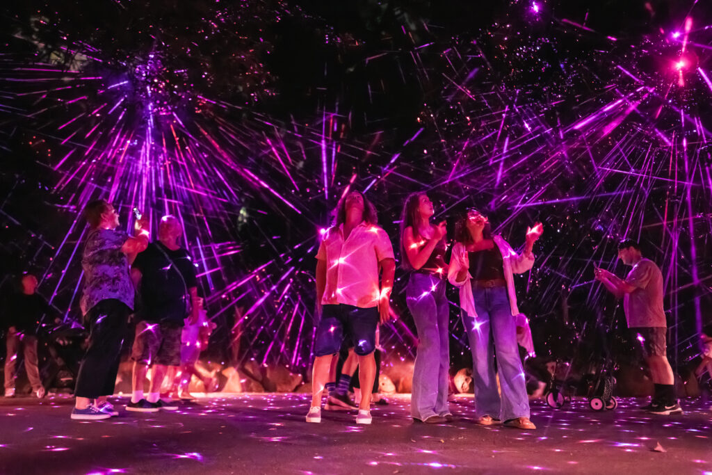 People walk through a wooded area illuminated by purple and pink laser lights, with beams shining through trees and lanterns lighting the path.