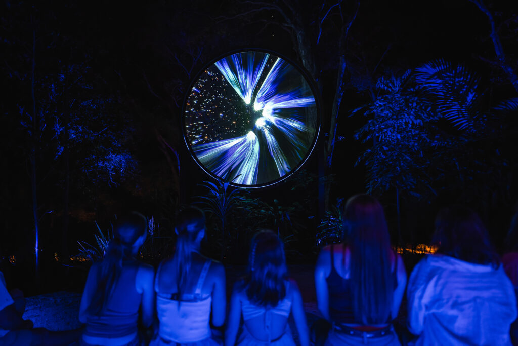 Five people sit in front of a large circular display with swirling blue lights, surrounded by illuminated trees at night.
