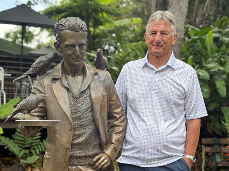 Man in shirt and shorts standing next to Alex Griffith statue looking alike, surrounded by flowers and trees