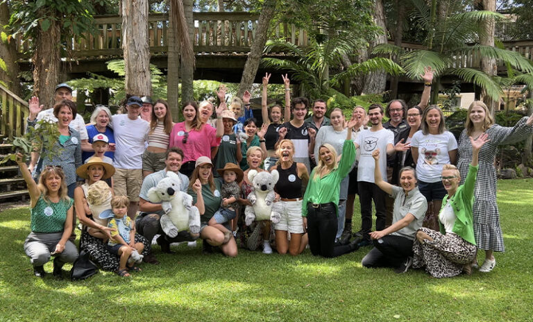 Group photo of people on green grass, hands up, some holding koala plush and eucalyptus branches, lush green trees in background