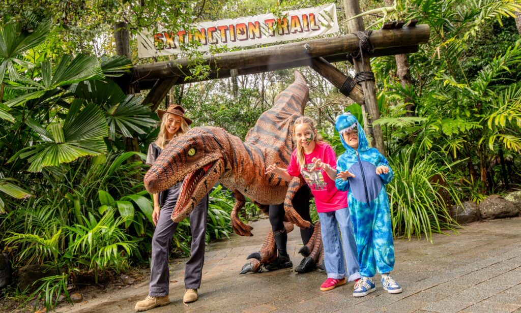 A girl in a pink dinosaur shirt and jeans and a boy in a blue dinosaur suit making raw gestures right next to a life-size dinosaur replica. A young woman wearing khaki outdoor clothing and a stetson hat is smiling standing on the other side of the dinosaur. They are in an outdoor area with tropical trees and a wooden arch with a sign saying extinction trail in the background