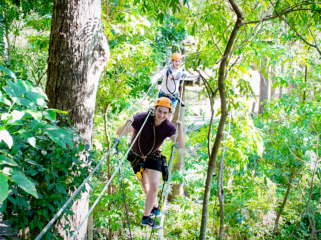 two young people wearing helmets and harnesses balance on a cable suspended in the air, through a dense forest with trees