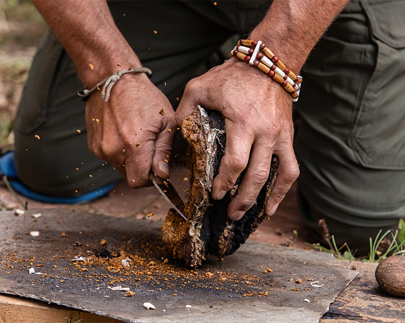 Man shaving wooden piece with a small blade, set on a sheet on the ground